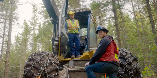 Two men operate heavy equipment in the forest.